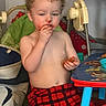 Ezra joined the competition — help win amazing prizes! toddler, child, curly_hair, plaid_pants, shirtless, snack, messy, high_chair, indoor, table, blue_table, red_pants, food, hand, face, nose, mouth, furniture, person, contemplative
