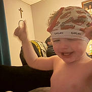 Ezra joined the competition — help win amazing prizes! toddler, child, headband, indoors, smiling, happy, blurry, bare_chested, arm_raised, living_room, crucifix, family_portrait, curtain, sofa, blanket, person, furniture, wall, ceiling, toy