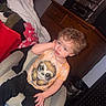 Ezra is registered to the contest to win money with this photo: toddler, child, curly_hair, orange_shirt, sloth_graphic, black_pants, chair, indoor, clutter, furniture, lamp, cabinet, clothing, blue_blanket, hand_on_face, curious_expression, carpet, trash_can, household_items, relaxed