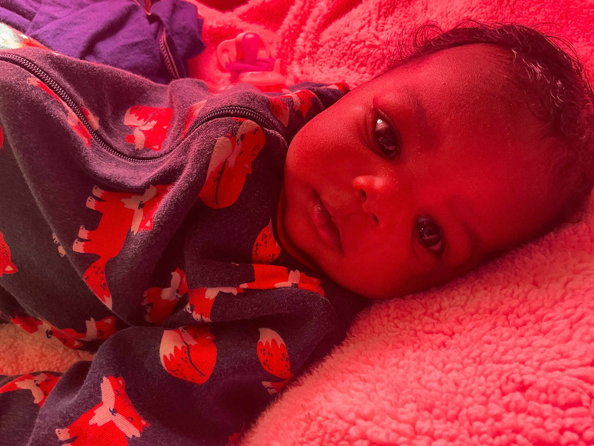 Amena is registered to the contest to win money with this photo: baby, infant, face, eyes, clothing, sleeper, fox_pattern, blanket, pink, soft_texture, lying_down, closeup, portrait, curious, indoors, child, cute, warm_light, cozy, sleepwear