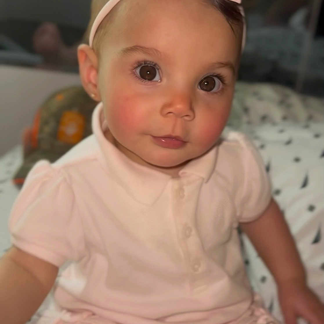 Kaila joined the competition — help win amazing prizes! toddler, child, baby, pink_bow, headband, pink_outfit, indoor, bedspread, patterned_bedspread, seated, curious_expression, brown_eyes, soft_lighting, portrait, young_child, cute, person, face, infant, clothing