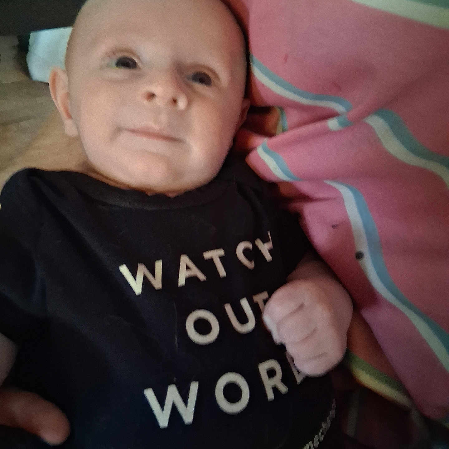 Raivo is registered to the contest to win money with this photo: baby, blanket, close_up, cuddle, cute, face, floor, hand, head, indoor, infant, newborn, onesie, parent_hand, pink_stripes, portrait, smile, soft_focus, text_on_clothing, tshirt