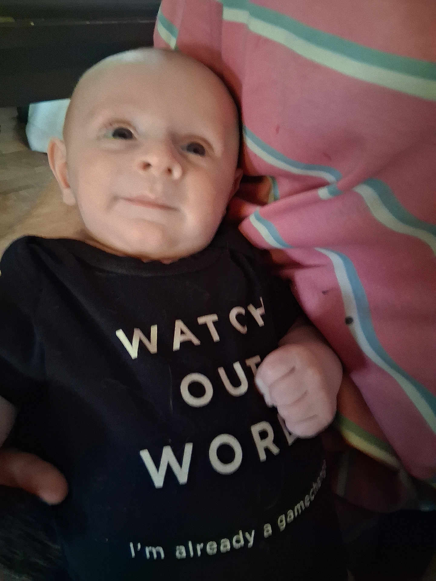 Raivo is registered to the contest to win money with this photo: baby, infant, newborn, onesie, text_on_clothing, smile, face, head, hand, close_up, portrait, indoor, blanket, pink_stripes, tshirt, cuddle, parent_hand, floor, cute, soft_focus