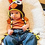 Weston joined the competition — help win amazing prizes! baby, infant, child, knitted_hat, turkey_hat, colorful, blue_jeans, brown_shirt, blanket, cozy, cute, portrait, sitting, indoor, soft_texture, young_child, headwear, clothing, person, expression