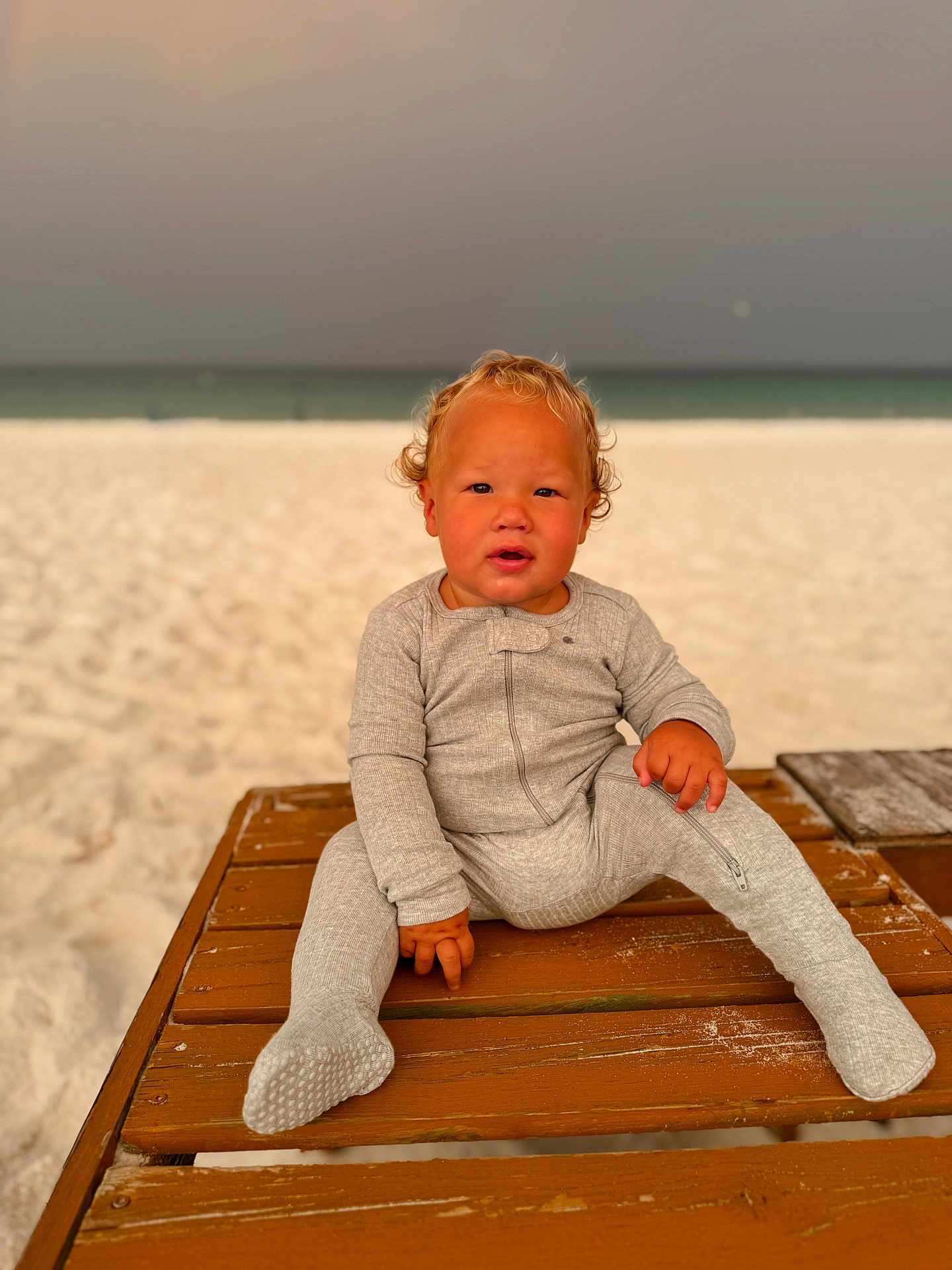 Landry joined the competition — help win amazing prizes! baby, beach, casual, child, curly_hair, cute, expression, grey_clothing, infant, onesie, outdoor, person, portrait, relaxed, sand, sea, sitting, sunset_lighting, toddler, wooden_bench