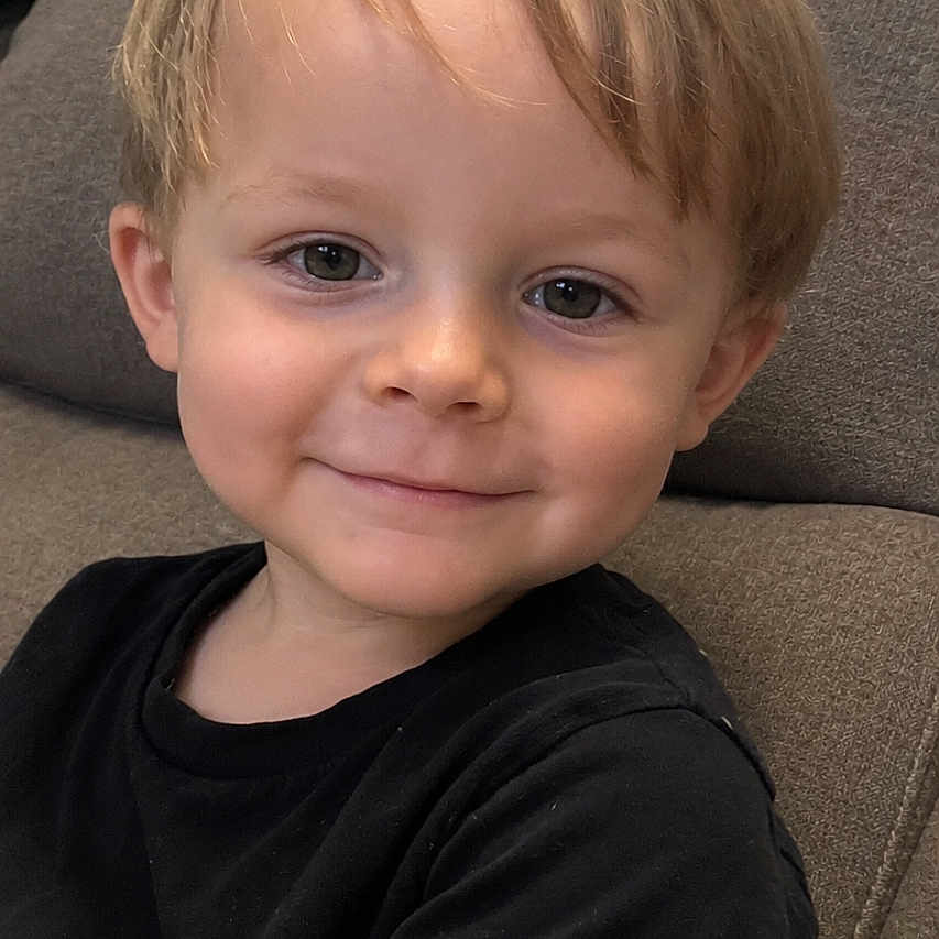 Oakley joined the competition — help win amazing prizes! child, toddler, portrait, smiling, happy, sitting, couch, indoor, black_shirt, blond_hair, brown_eyes, face, head, arm, casual, home, furniture, close_up, photography, adorable