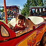 amusement_ride, carnival, child, daytime, fair, fun, game_stalls, happy, number_95, outdoor, person, playful, plush_toys, red, smiling, sunlight, toddler, toy_car, trees, yellow
