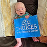 baby, infant, sign, blue_sign, text, couch, leather, pillow, knitted_pillow, yellow_pillow, white_pillow, hand, foot, person, indoor, relaxed, sitting, child, face, holding