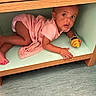 toddler, child, pink_clothing, toy, croissant_toy, wooden_table, cubbyhole, floor, indoor, curious, hiding, playful, small_space, barefoot, infant, person, young_child, looking_up, crawling, home