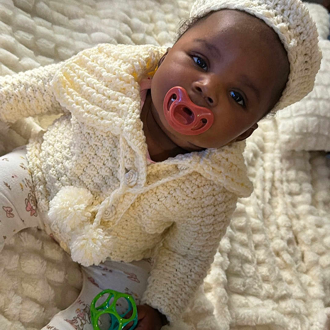 Knowledge is registered to the contest to win money with this photo: baby, infant, pacifier, knitwear, hat, blanket, toy, hanger, cute, child, indoors, portrait, sitting, soft_texture, warm_clothing, comfort, playful, cozy, person, young_child