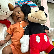 Rugvednandan is registered to the contest to win money with this photo: baby, plush_toy, mickey_mouse, white_bear, orange_outfit, bracelet, striped_blanket, lying_down, indoor, soft_toys, child, cute, cozy, floor, playtime, smiling, colorful, stuffed_animal, person, infant