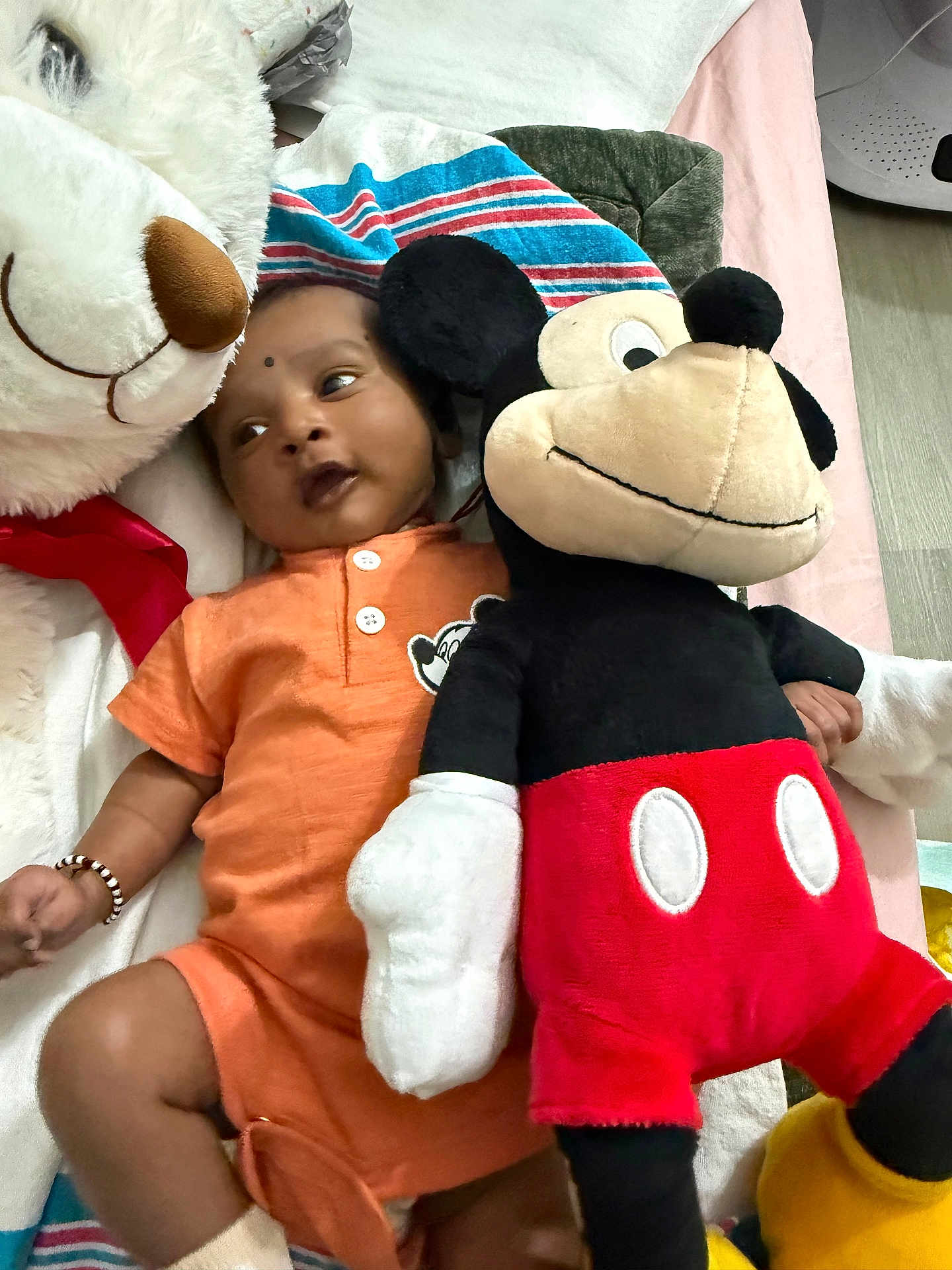 Rugvednandan is registered to the contest to win money with this photo: baby, plush_toy, mickey_mouse, white_bear, orange_outfit, bracelet, striped_blanket, lying_down, indoor, soft_toys, child, cute, cozy, floor, playtime, smiling, colorful, stuffed_animal, person, infant