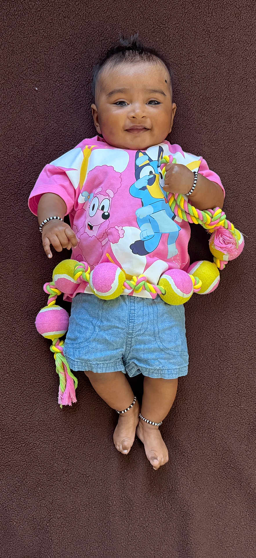 Rugvednandan is registered to the contest to win money with this photo: child, baby, infant, smiling, face, eyes_open, tshirt, denim_shorts, bracelet, anklet, toy, rope_toy, tennis_balls, colorful, lying_down, blanket, portrait, cute, hair, skin