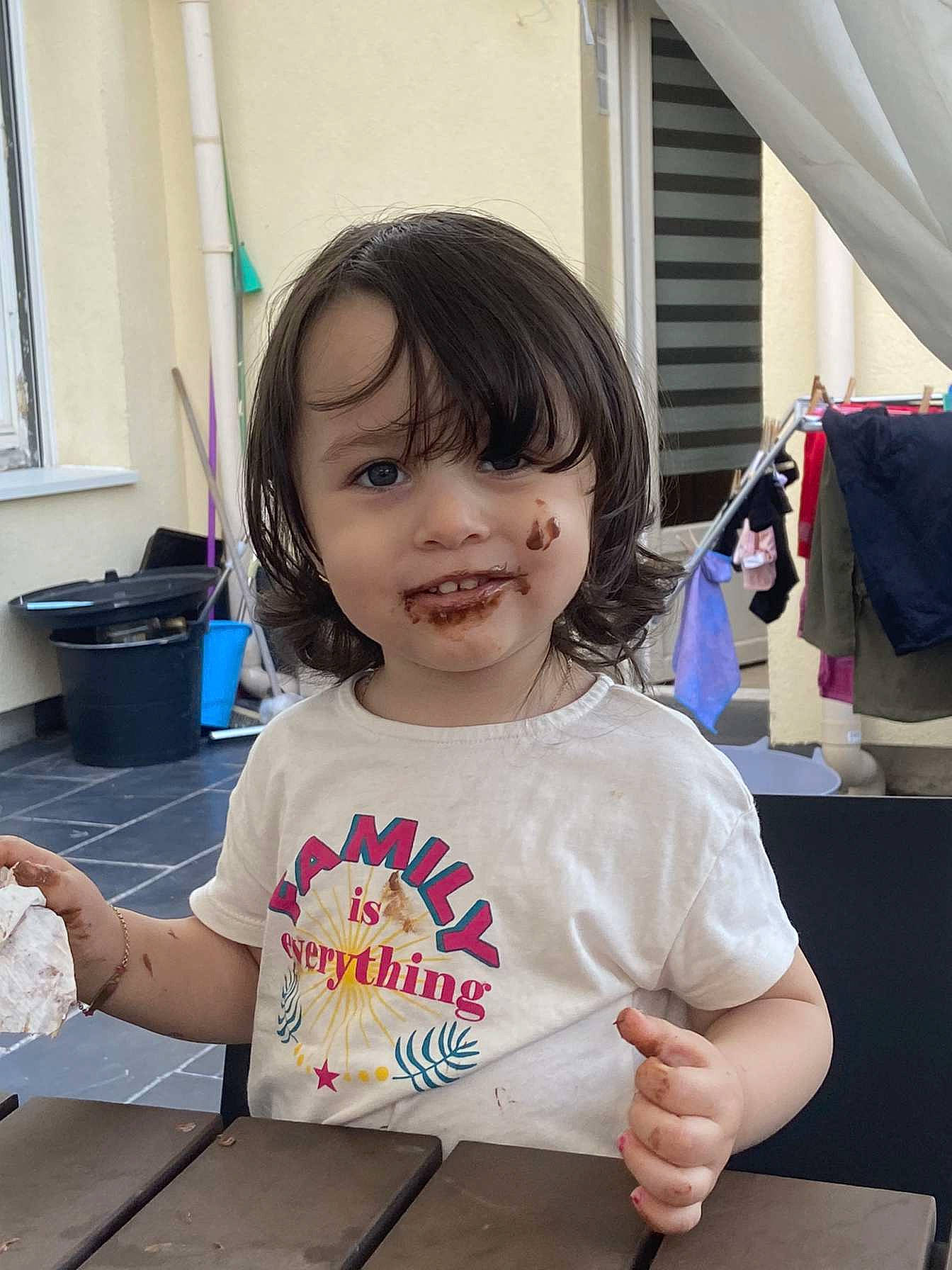 Inaya participe au concours pour gagner de l'argent avec cette photo : bag, bangs, chair, child, curtain, fun, happy, leisure, luggage_and_bags, person, room, sitting, sleeve, smile, t_shirt, table, toddler, vacation, window