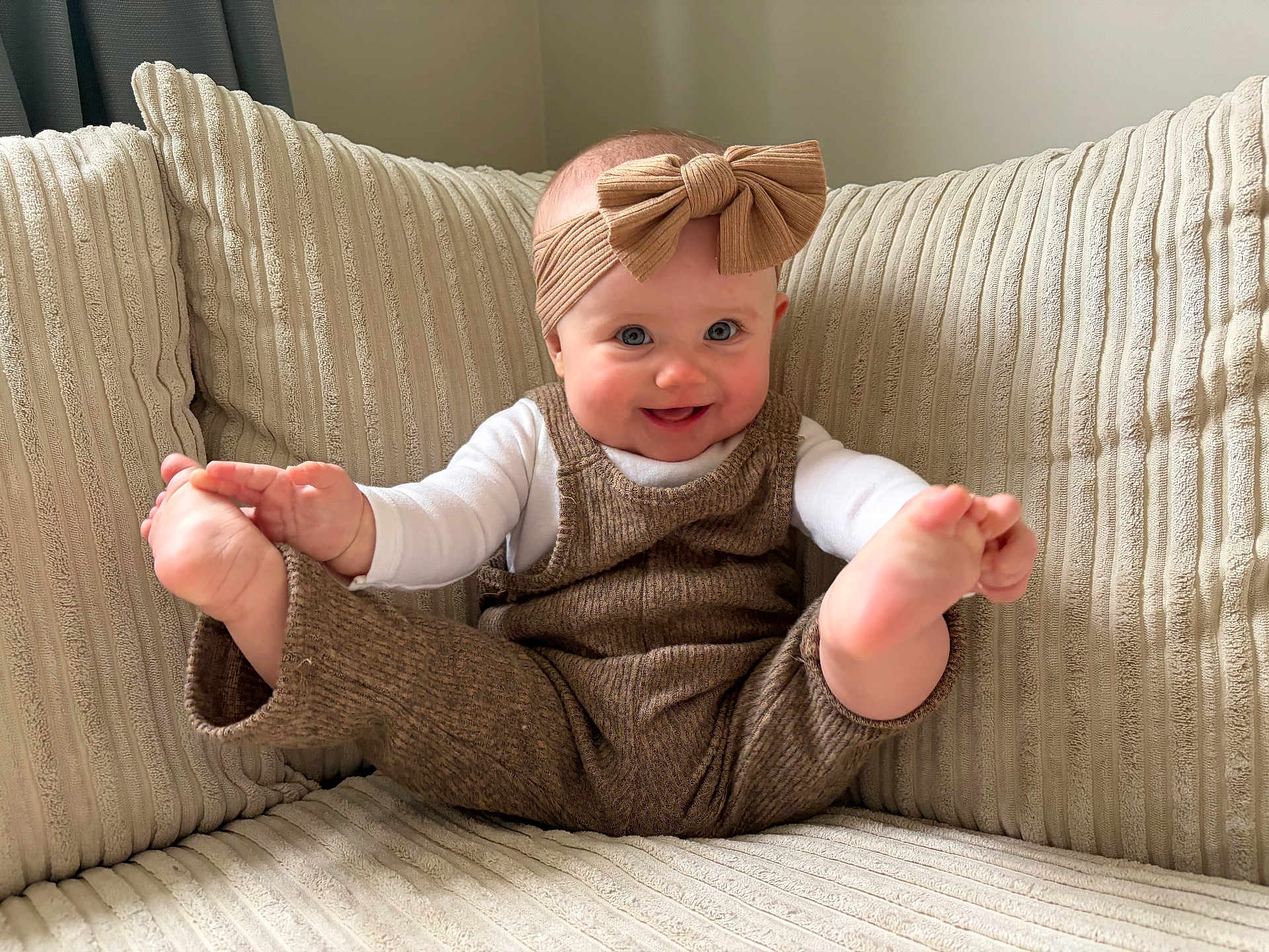 Paisley joined the competition — help win amazing prizes! baby, child, smiling, headband, bow, overalls, couch, beige, texture, happy, playful, feet, hands, indoors, person, infant, cute, cozy, clothing, portrait