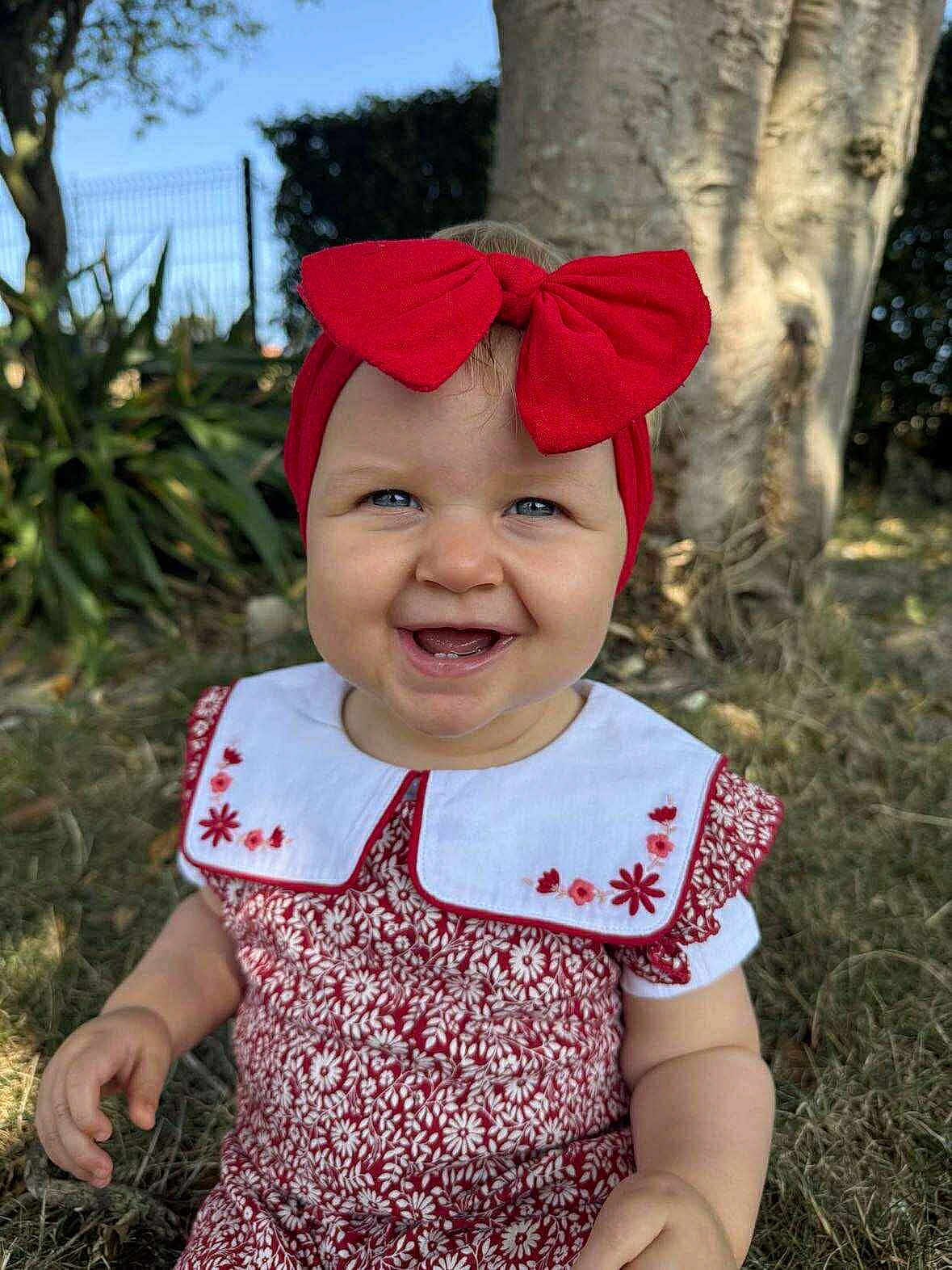 Lou a rejoint le concours — aidez-le/la à gagner de superbes lots ! baby, child, smile, red_bow, headband, dress, floral_pattern, outdoor, grass, tree, sunlight, blue_eyes, cute, happy, portrait, nature, embroidery, sitting, toddler, person