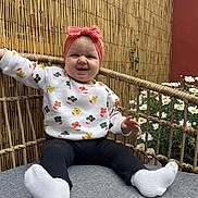 Lou a rejoint le concours — aidez-le/la à gagner de superbes lots ! baby, child, smiling, headband, pink_bow, white_socks, black_pants, floral_sweater, cushion, gray_cushion, bamboo_fence, daisies, flowers, outdoor, garden, happy, person, sitting, cute, portrait