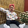 baby, child, smiling, headband, pink_bow, white_socks, black_pants, floral_sweater, cushion, gray_cushion, bamboo_fence, daisies, flowers, outdoor, garden, happy, person, sitting, cute, portrait