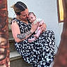 affection, baby, baby_clothes, casual_clothing, closeup, emotion, face, holding, love, natural_light, outdoor, patterned_dress, person, portrait, sitting, skin, staircase, stairs, tattoo, woman