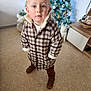 brown_shoes, cabinet, checkered, child, christmas_tree, coat, curious, curly_hair, festive, floor, furniture, holiday_decorations, indoor, lights, ornaments, portrait, standing, toddler, toy, winter_clothing