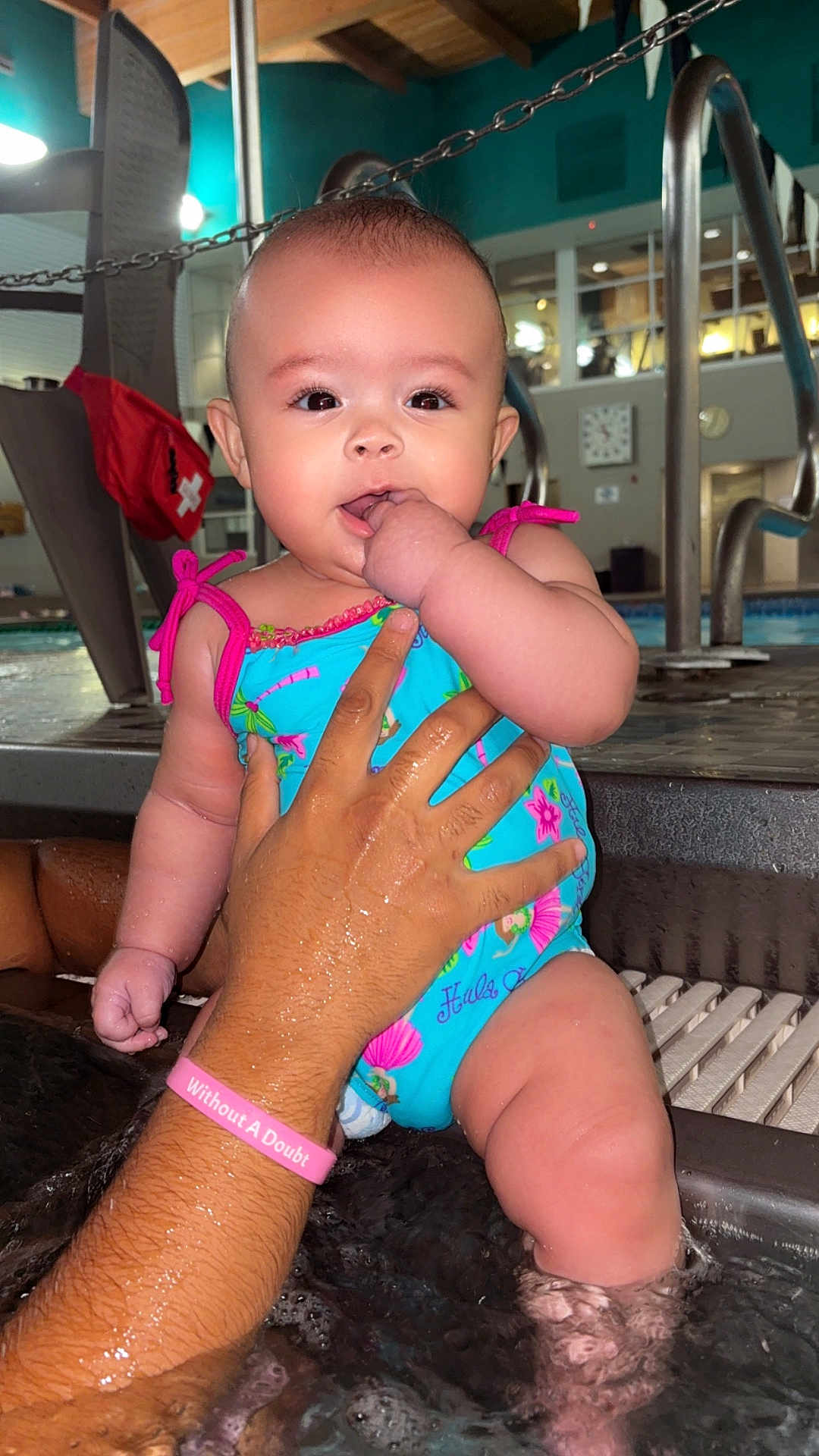 Laylani Dailynn joined the competition — help win amazing prizes! baby, child, swimsuit, pool, water, hand, adult_hand, face, indoor, chair, clock, person, skin, water_splash, colorful_clothing, support, infant, curious, wet, playful