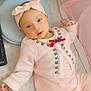baby, infant, pink_clothing, headband, bow, crib, blanket, remote_control, patterned_sheet, lying_down, cute, soft_lighting, indoor, child, sleepy, cozy, portrait, person, newborn, resting