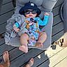 baby, child, sunglasses, bucket_hat, mickey_mouse, outdoor, deck, wooden_floor, tattoo, barefoot, cushion, chair, clothing, person, relaxing, summer, infant, cute, fashion, sunlight