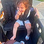 toddler, child, stroller, basketball, toy, footwear, outdoor, sidewalk, grass, sunlight, playing, hand, clothing, seat_belt, happy, person, summer, shorts, shoe, hair