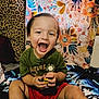 toddler, child, smiling, laughing, toy, green_shirt, red_shorts, blanket, floral_pattern, leopard_print, indoor, happy, cute, sitting, playful, person, face, head, legs, colorful
