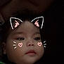 baby, child, face, cat_ears_filter, digital_filter, heart_cheeks, dark_background, cartoon_print, clothing, cute, portrait, indoors, young_child, expressionless, whiskers, head, closeup, soft_light, casual, person