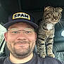 Spud is registered to the contest to win money with this photo: man, cat, tabby_cat, glasses, hat, cap, smile, beard, mustache, indoor, car, seat, shoulder, pet, animal, companion, curious, wide_eyes, casual_clothing, friendship