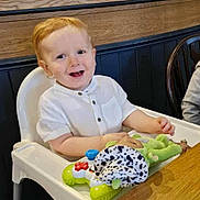 Elliot joined the competition — help win amazing prizes! toddler, child, high_chair, toy, smiling, indoor, table, wood, white_clothing, red_hair, happy, person, playful, seat, furniture, cute, face, young_child, casual, baby