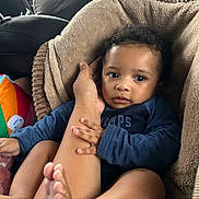 Samuel is registered to the contest to win money with this photo: baby, infant, child, portrait, car, car_seat, blanket, adult_hand, family, cozy, sitting, lying_down, wide_eyed, curly_hair, blue_shirt, colorful_toy, tiny_feet, indoor, vehicle, adorable