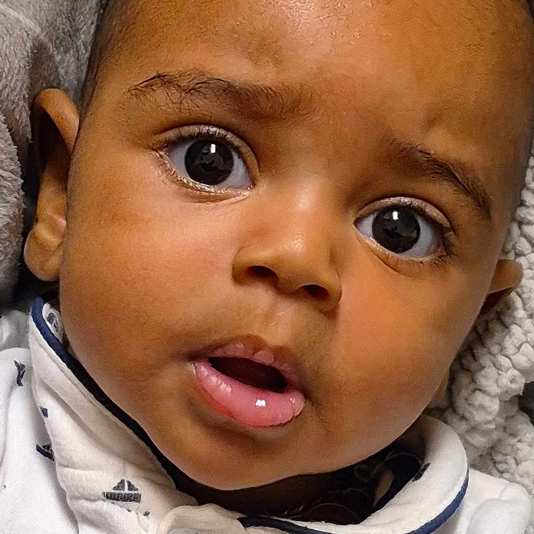 Aisaiah is registered to the contest to win money with this photo: baby, blanket, child, close_up, clothing, cozy, curious, cute, expression, eyes, face, hands, indoors, infant, mouth, portrait, sailboat_pattern, skin, white_shirt, young