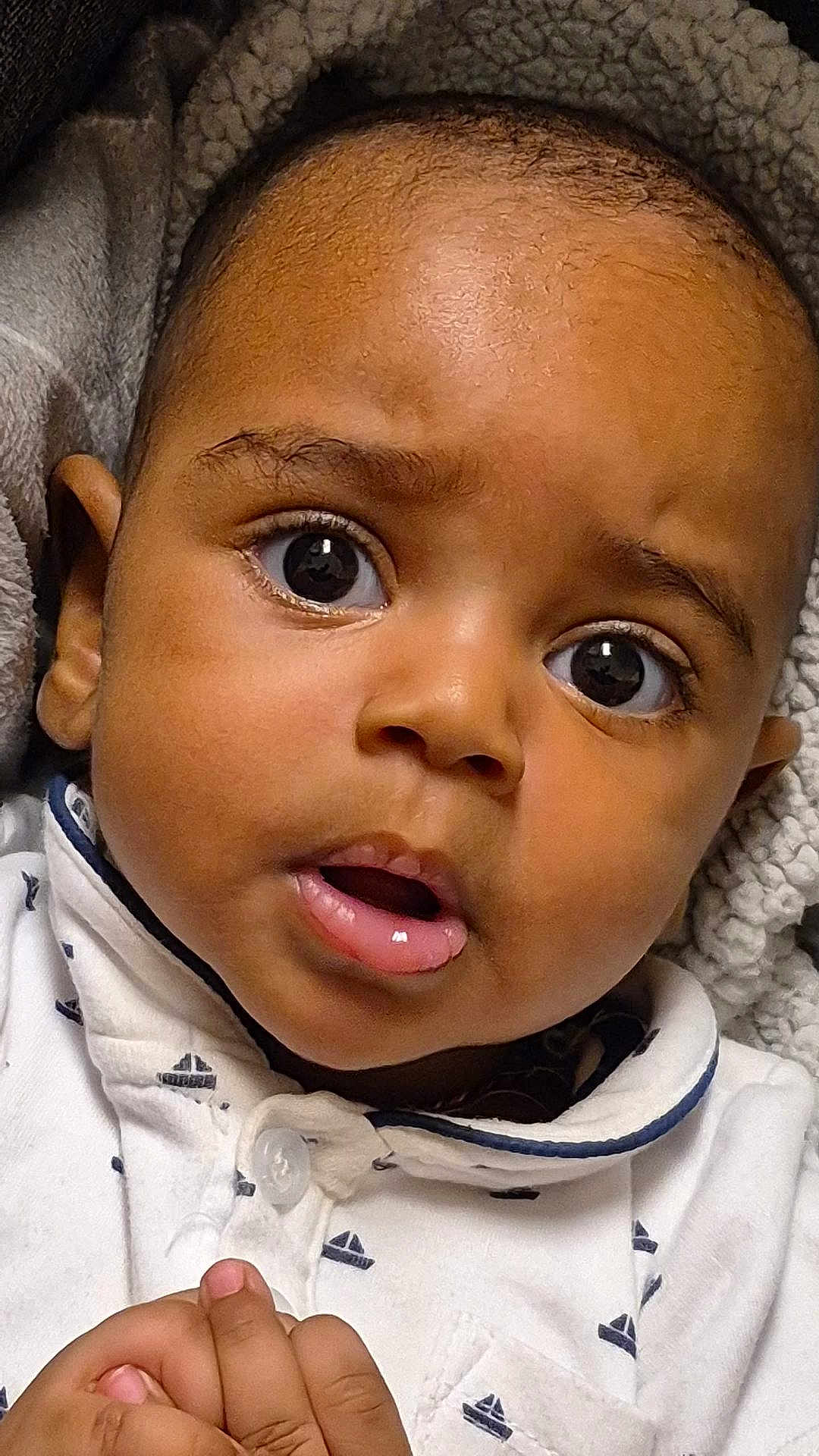 Aisaiah is registered to the contest to win money with this photo: baby, close_up, face, curious, expression, infant, skin, eyes, mouth, hands, blanket, clothing, white_shirt, sailboat_pattern, cute, indoors, portrait, child, young, cozy