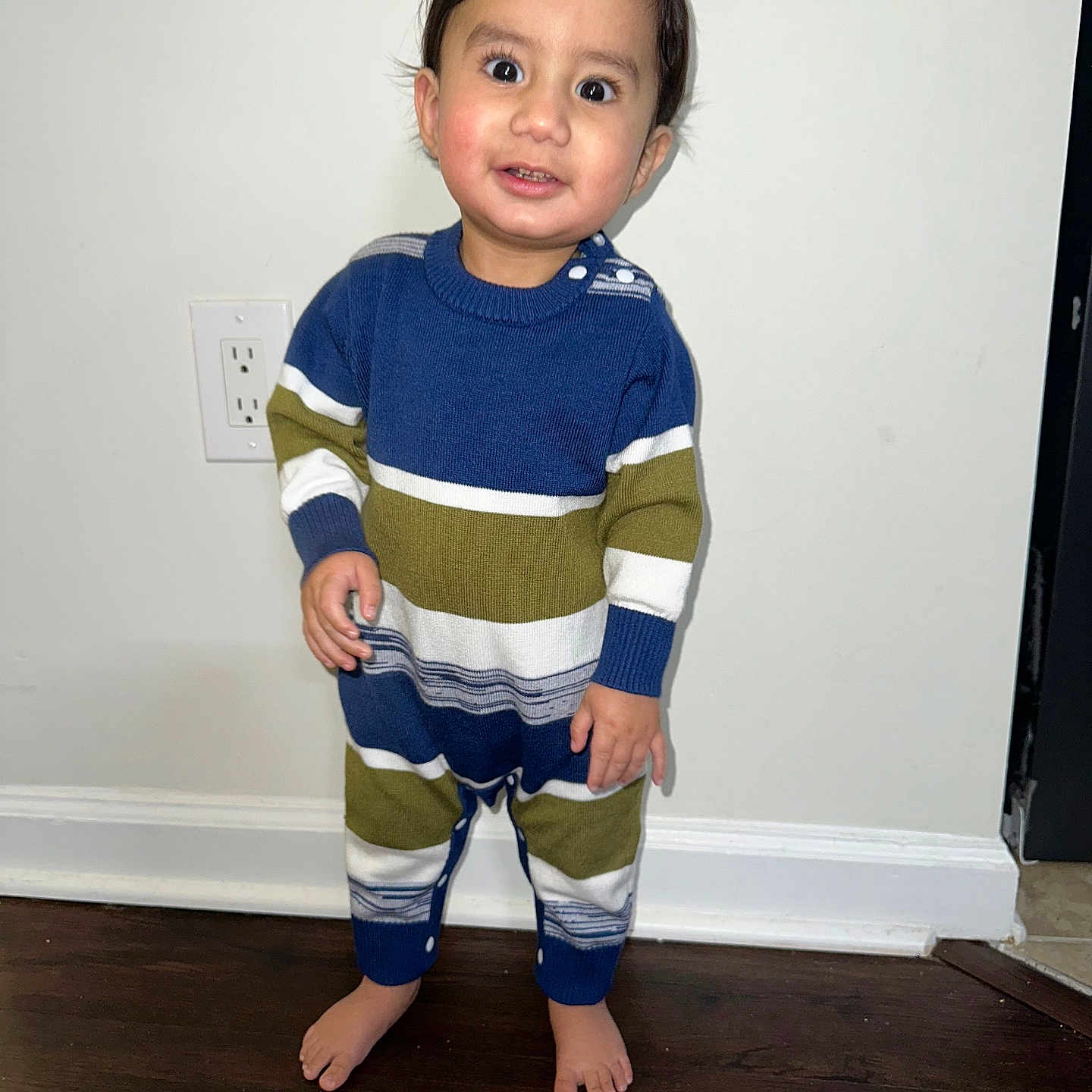 Kavir joined the competition — help win amazing prizes! bare_feet, barefoot, buttoned_onesie, child, cozy_clothing, curious, cute, flooring, indoor, long_sleeves, person, portrait, power_outlet, smiling, standing, striped_onesie, toddler, white_wall, wooden_floor, young_child