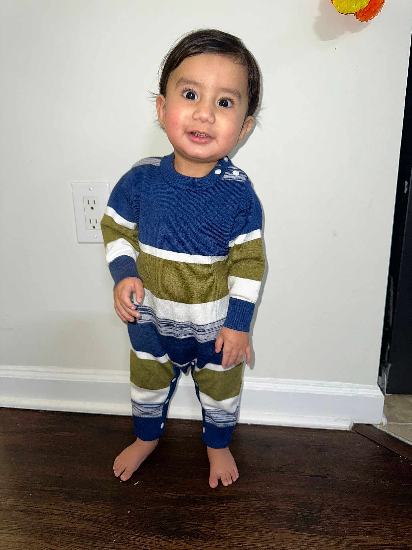 Kavir joined the competition — help win amazing prizes! toddler, child, barefoot, striped_onesie, indoor, wooden_floor, white_wall, power_outlet, smiling, curious, standing, young_child, person, portrait, cozy_clothing, long_sleeves, buttoned_onesie, flooring, bare_feet, cute