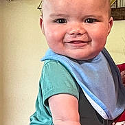 Iker is registered to the contest to win money with this photo: baby, child, smiling, blue_bib, green_shirt, adult_hand, indoor, face, portrait, holding, red_shirt, decor, anchor, wooden_anchor, model_ship, furniture, cute, happy, skin, person
