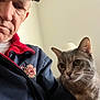 man, cat, indoor, portrait, pet, animal, person, face, clothing, cap, jacket, gray_cat, companion, calm, closeup, casual, neutral_background, indoor_lighting, expression, senior