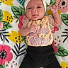 baby, black_skirt, bow, child, closeup, clothing, colorful, cute, dress, face, floral_blanket, headband, indoors, infant, lying_down, patterned_outfit, photography, portrait, raised_hand, smiling