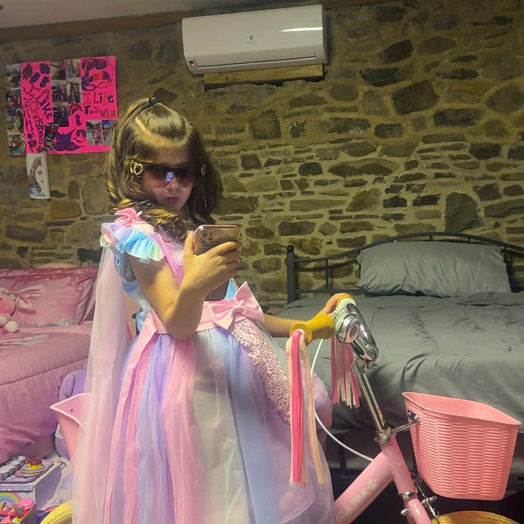 Shpresa is registered to the contest to win money with this photo: child, girl, bicycle, pink, dress, sunglasses, indoor, bed, stone_wall, training_wheels, toy, footwear, sock, cape, phone, bedroom, poster, decor, furniture, playful