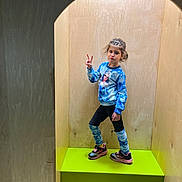 Shpresa is registered to the contest to win money with this photo: child, girl, pose, peace_sign, blue_sweater, character_print, leg_warmers, sneakers, pink, black, green_platform, wooden_walls, indoor, fashion, portrait, standing, playful, expression, casual_clothing, minimalistic