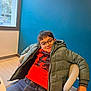 child, boy, glasses, green_jacket, red_sweater, jeans, chair, indoor, window, blue_wall, carpet, casual, relaxed, portrait, seated, modern, office, smile, person, young