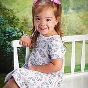 Savannah is registered to the contest to win money with this photo: child, girl, smile, pink_bows, dress, white_bench, sitting, portrait, happy, outdoor_background, greenery, flower_pattern, cute, young, face, human, person, hair, dimples, casual