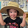 toddler, child, hat, straw_hat, shopping_cart, black_tshirt, flowers, text, rosy_cheeks, smiling, curious_eyes, indoor, store, aisle, holding_hat, cute, happy, portrait, short_hair, casual_clothing