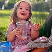 Savannah is registered to the contest to win money with this photo: child, girl, smile, happy, outdoor, daylight, plastic_cup, pink_dress, animal_print, chair, grass, sunlight, nature, face, person, casual, sitting, playful, joyful, relaxed