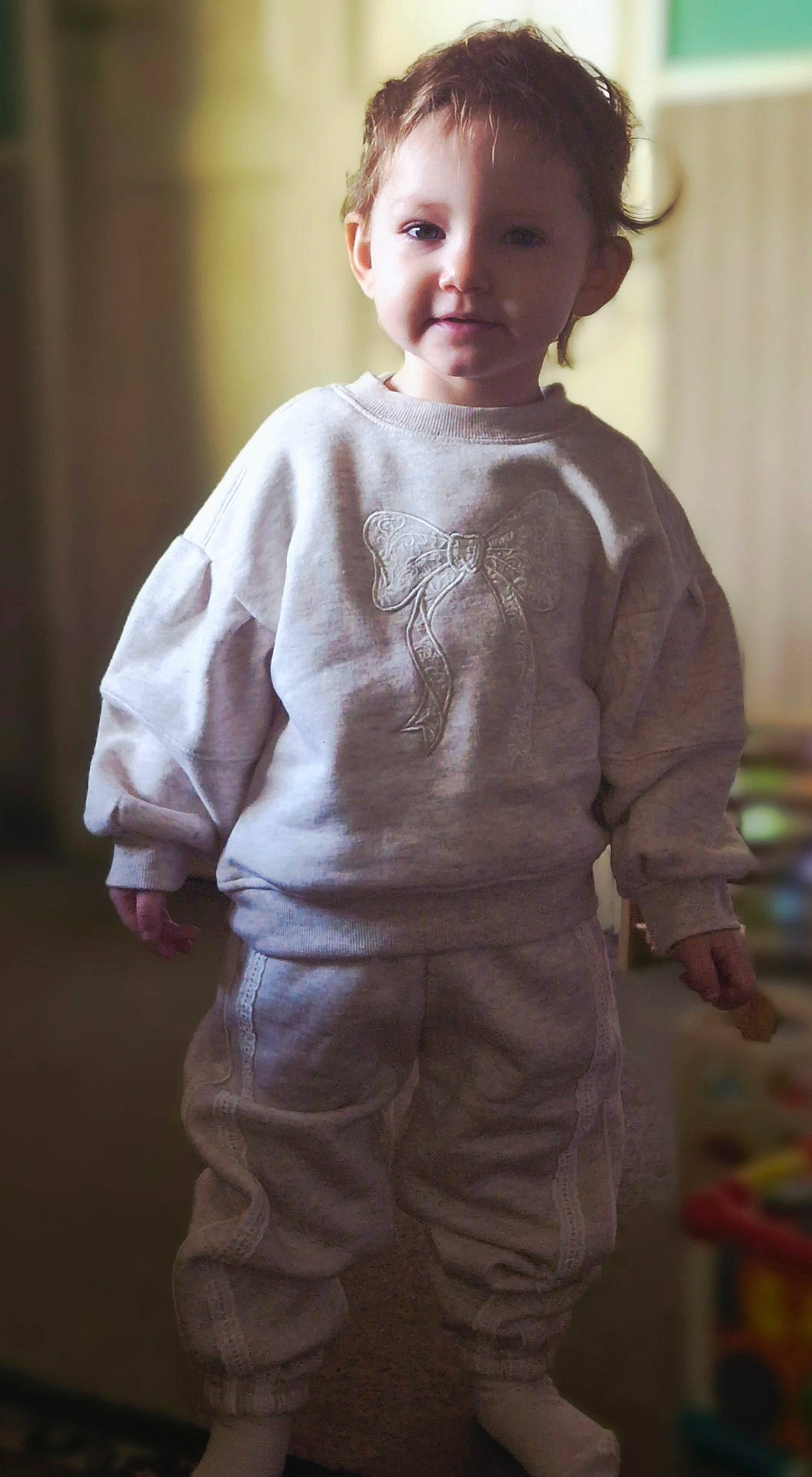 Skylar-Rose is registered to the contest to win money with this photo: blurred_background, bow_design, casual, child, clothing, cozy, face, hair, home, indoor, light, person, portrait, smile, socks, standing, sweatpants, sweatshirt, toddler, young_child