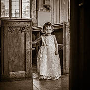 Skylar-Rose joined the competition — help win amazing prizes! child, girl, dress, church, wooden_pews, window, light, stone_floor, cross_necklace, interior, vintage, curly_hair, standing, portrait, sepia, religious, architecture, young, solemn, decorative