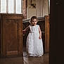 child, toddler, white_dress, church, pew, wooden_furniture, natural_light, window, floor, portrait, indoor, ceremony, cute, girl, floor_tiles, cross_necklace, curly_hair, standing, dress, solemn