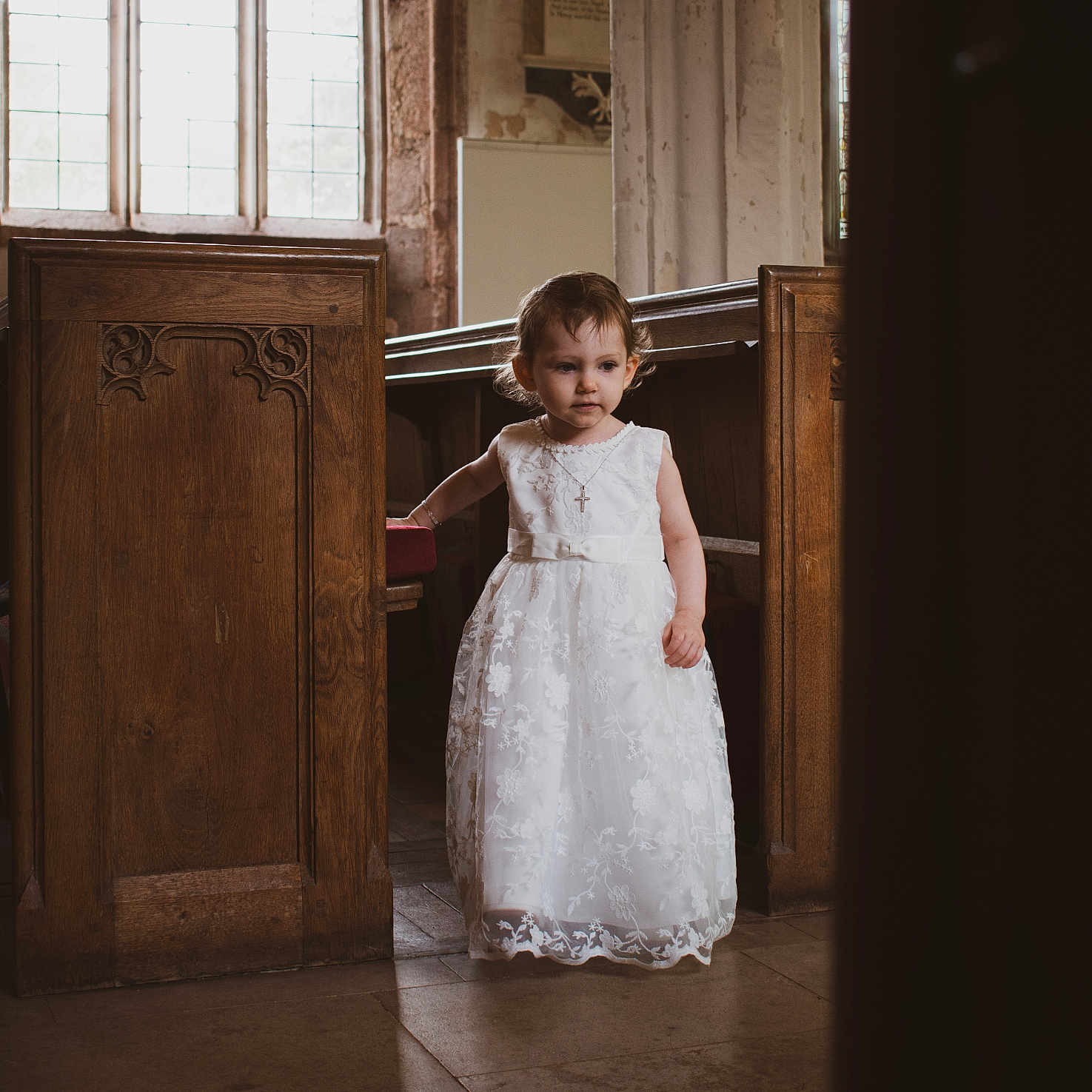 Skylar-Rose joined the competition — help win amazing prizes! ceremony, child, church, cross_necklace, curly_hair, cute, dress, floor, floor_tiles, girl, indoor, natural_light, pew, portrait, solemn, standing, toddler, white_dress, window, wooden_furniture